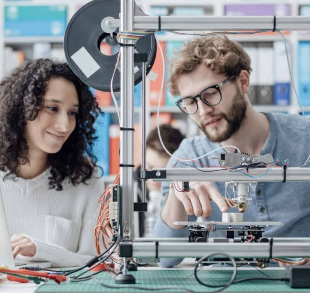 Two people collaborate on a 3D printer, examining its components and wiring in a brightly lit workspace during a 2023 Hackathon.