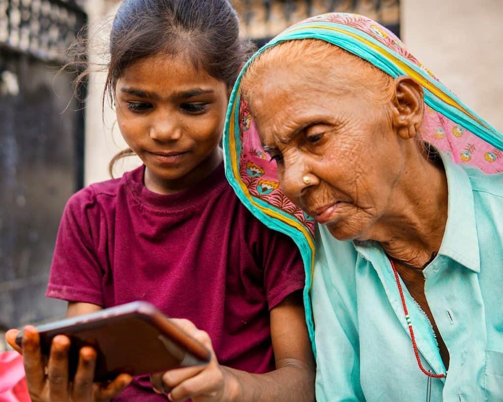 A young girl in a maroon shirt shows a smartphone to an elderly woman wearing a colorful headscarf and teal blouse. They sit closely, focused on the screen, as digital technologies spark moments of connection and women empowerment between generations.