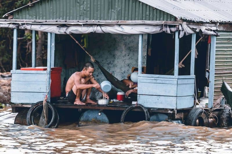 Amidst the murky waters, a man pours water into a pot on the porch of an informal floating house. Inside, another person reclines, embodying the quiet resilience often seen in urban areas facing infrastructure challenges.