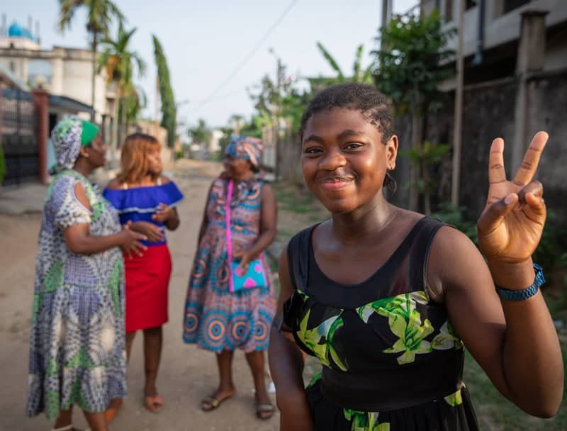 Young girl smiling and making a peace sign in the foreground with three empowering women discussing digital technologies in the background on a sunny street.