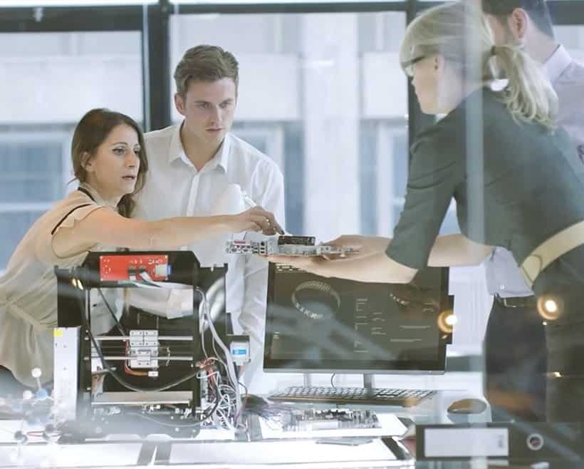 Four people in an office collaborate on an electronics project, using a 3D printer and computer with 3DEXPERIENCE by Dassault Systèmes to streamline product lifecycle management as one person passes a circuit board to another.