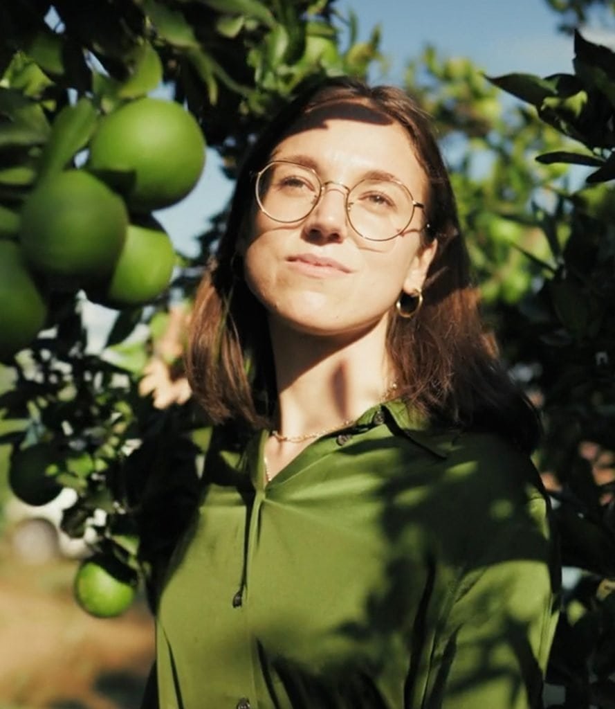 Standing among the lush citrus trees of Agrumaria Reggina, a woman in glasses and a green shirt gazes upward.