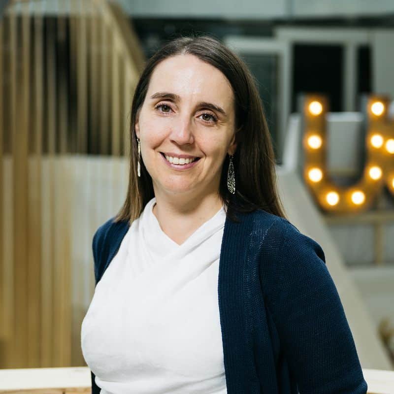 A woman with long dark hair, wearing a white blouse and dark cardigan, stands indoors, smiling at the camera. There's a lit-up letter "U" in the background.