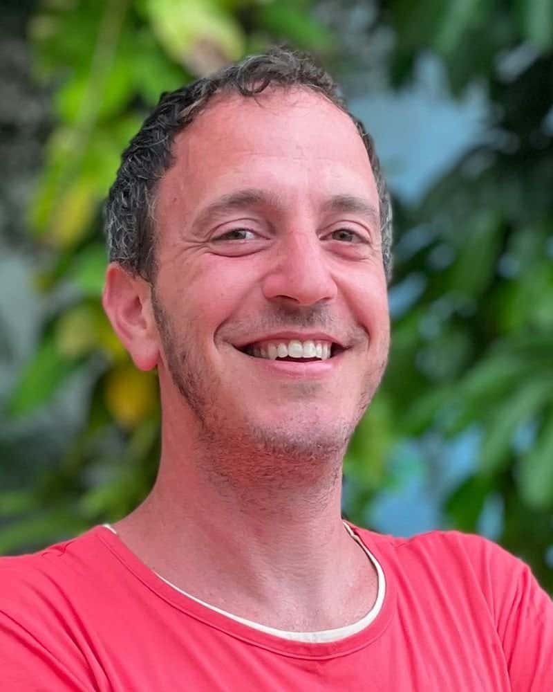 Bryce Neuman, in a red shirt, stands smiling against a leafy green background.