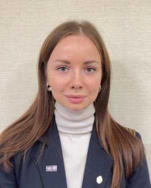 Dr. Valeriia Starshaia, a woman with long brown hair, stands confidently against a plain background, wearing a black blazer and white turtleneck as she looks directly at the camera.
