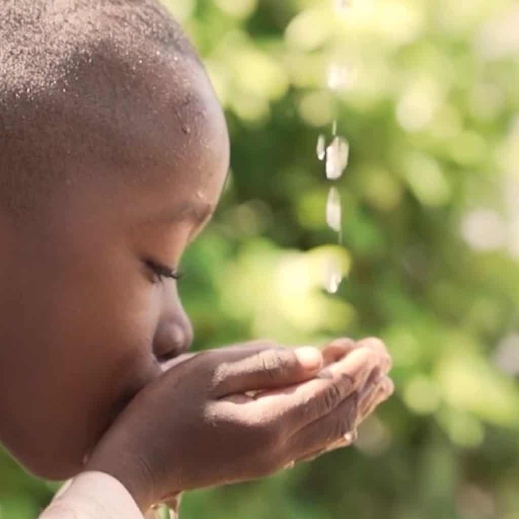 Child drinking water from cupped hands, with water streaming down. Bright, blurred foliage in the background.