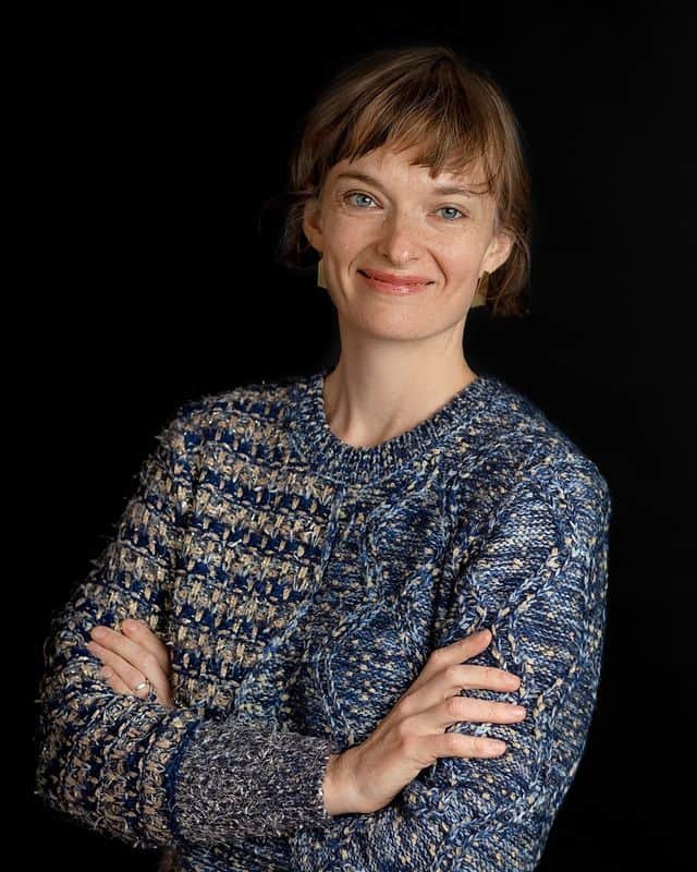 Dr. Eleonore Fournier-Tombs, with short hair, smiles confidently in a blue patterned sweater, crossing her arms against a black background.