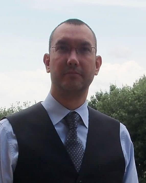 Dr. Rodrigo Perez Fernandez, wearing glasses, a blue shirt, a tie, and a black vest, stands outdoors with trees and a cloudy sky in the background.
