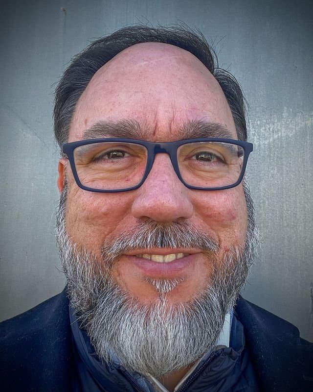 Ramon Manresa, a man with glasses and a graying beard, smiles slightly while looking directly at the camera. He dons a dark jacket against a plain gray background.