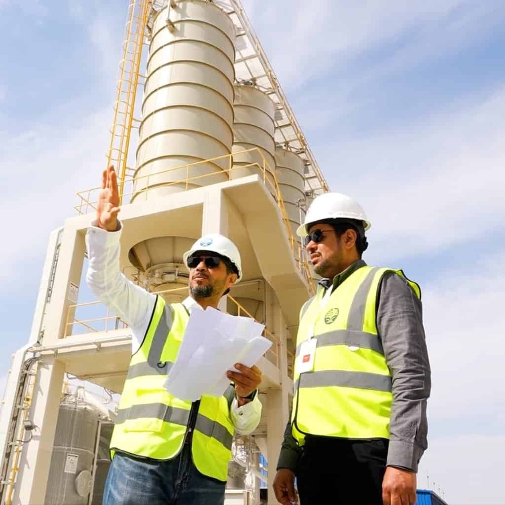 Two men in safety vests and helmets stand in front of an industrial structure. One holds papers and gestures upward while the other listens attentively, a scene typical of a Saudi Water Partnership Company project. Bright daylight illuminates the setting.