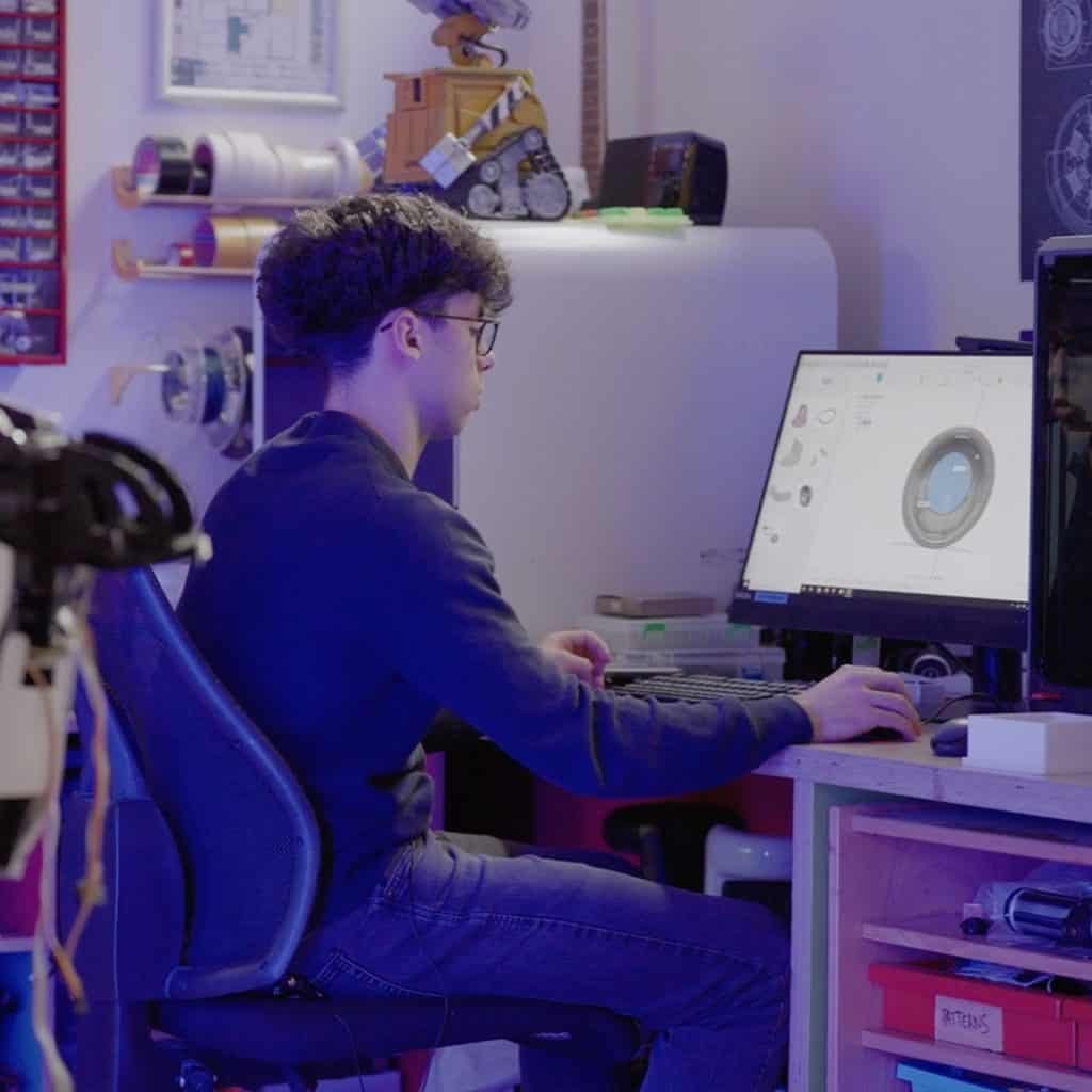 In a room filled with technical equipment and shelves, a person in glasses intently works on a 3D design displayed on their computer screen, honing skills that reflect the innovative spirit found at an international school in Geneva.