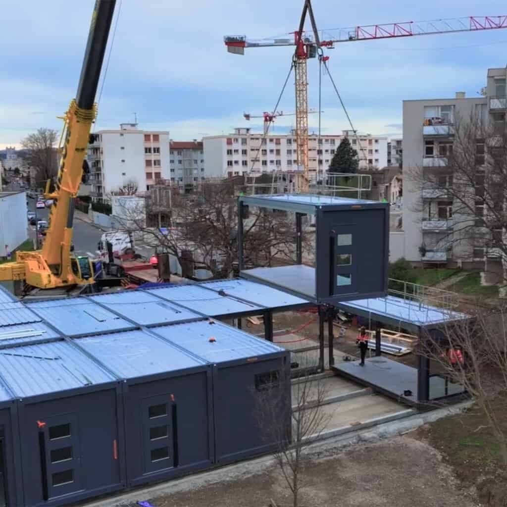 A crane lifts a modular building unit from Modulaire Group into place at a construction site in an urban residential area with apartment buildings in the background.