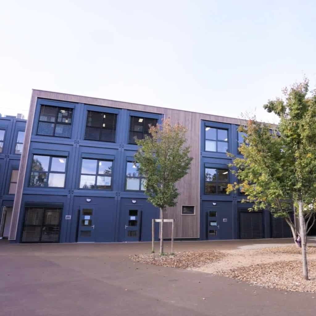 A three-story modern Modulaire Group building with blue and wood paneling, large windows, and two trees in front on a paved courtyard.
