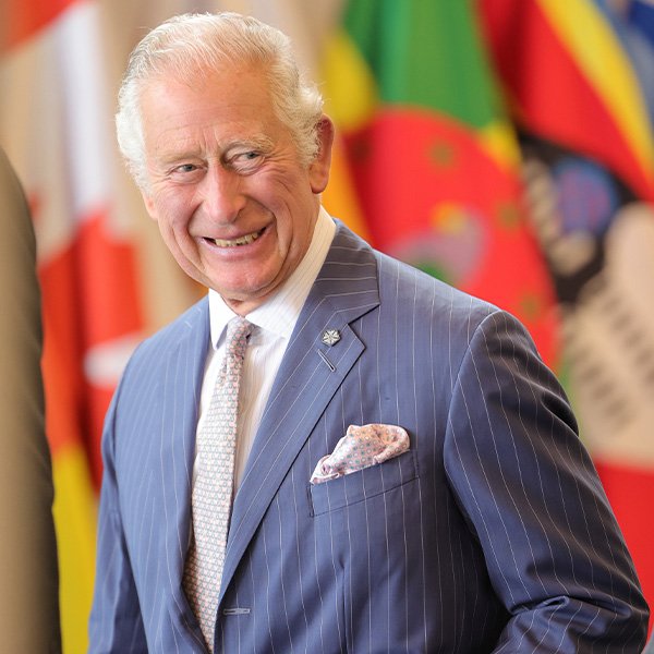 An older man in a blue pinstripe suit stands and smiles, with colorful international flags blurred in the background, reflecting the unity and diversity celebrated during the Commonwealth at 75.