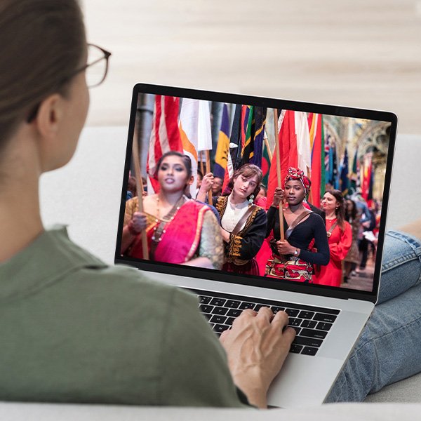 A person watches a laptop screen displaying a parade with people in traditional clothing holding various national flags, celebrating the Commonwealth at 75.