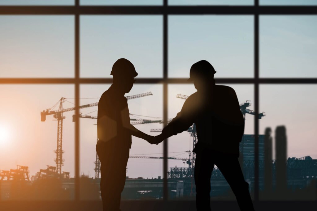 Two people wearing hard hats shake hands indoors with large windows overlooking construction cranes at sunset.