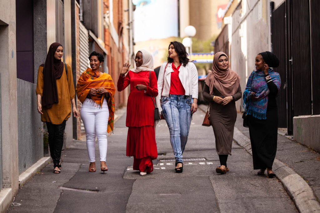 Six women walk together down an urban alleyway, engaged in conversation, wearing a variety of colorful outfits and headscarves.