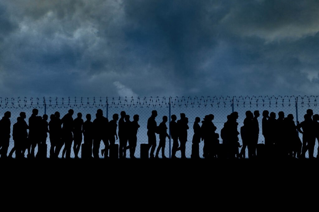 Silhouettes of people walking and carrying belongings in front of a barbed wire fence under a cloudy sky.