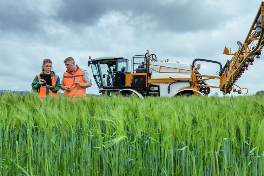 Two people in safety vests examine a tablet in a green crop field next to a large agricultural machine under a cloudy sky.