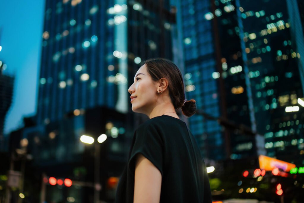 Woman in black top stands outdoors at dusk, with tall glass buildings and city lights in the background.