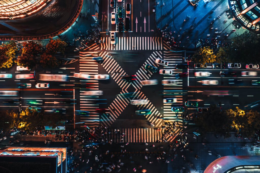 Aerial view of a busy city intersection at night with moving cars, crosswalks, and blurred lights, surrounded by buildings and crowds of people.