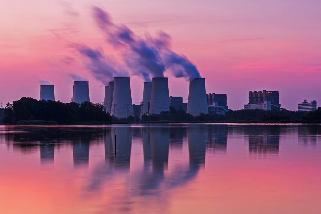 Cooling towers of a power plant emit steam at sunset, reflected in a calm body of water, with buildings and trees in the background.