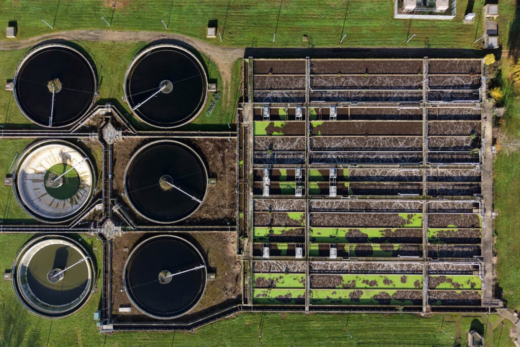 Aerial view of a wastewater treatment plant showing circular settling tanks on the left and rectangular filtration pools on the right, surrounded by green grass.