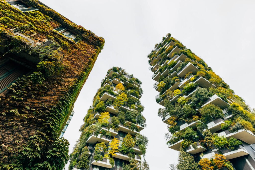 Three tall buildings covered with greenery and plants, featuring balconies filled with trees and shrubs, set against a cloudy sky.