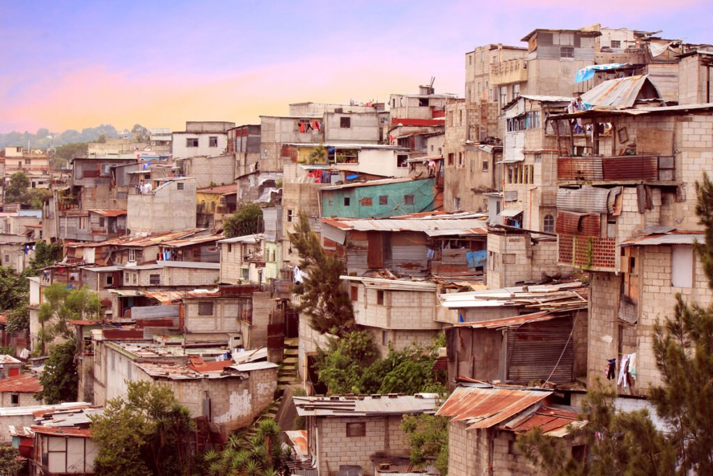 Crowded hillside community with densely packed concrete and metal sheet houses under a colorful sky.