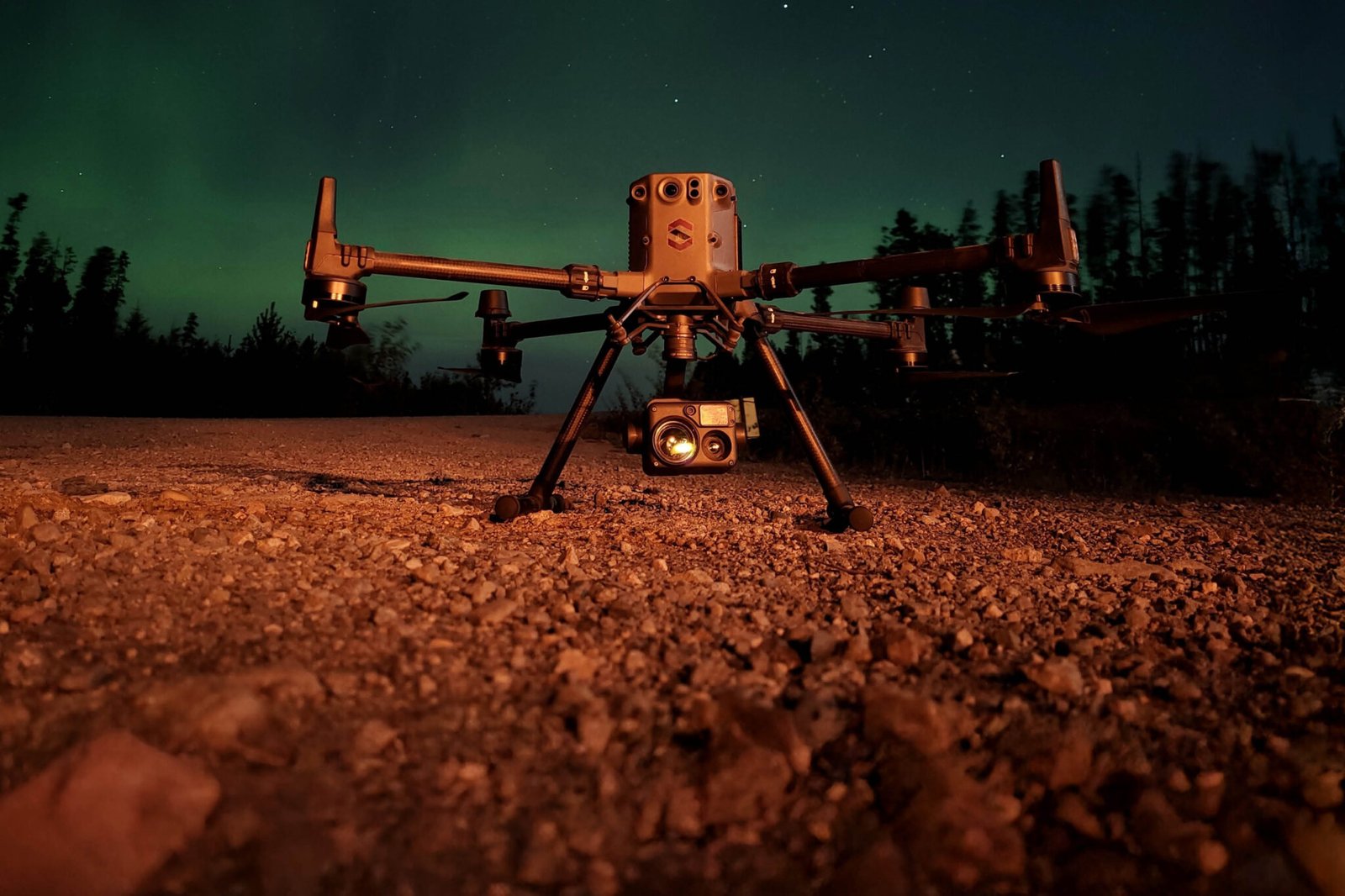 A drone with mounted camera is positioned on a gravel surface at night, illuminated by artificial light, with trees and a starry sky in the background.