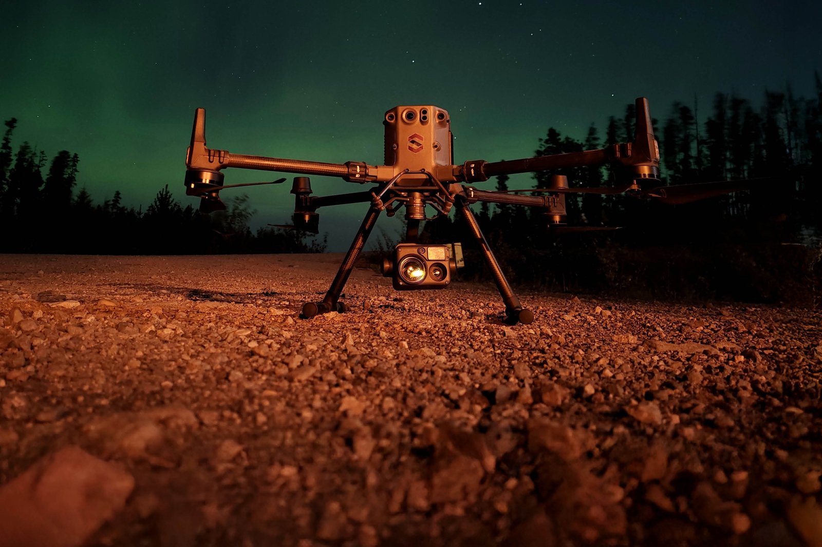 A drone with mounted camera is positioned on a gravel surface at night, illuminated by artificial light, with trees and a starry sky in the background.