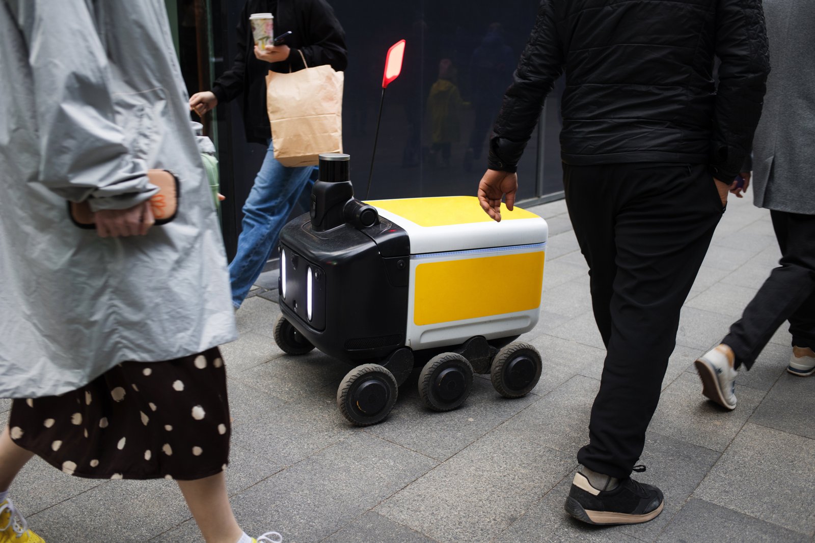 A small six-wheeled delivery robot moves along a city pavement among pedestrians