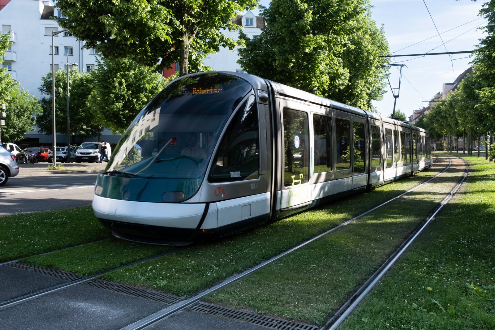 A modern tram travels on grass-covered tracks beside a tree-lined city road